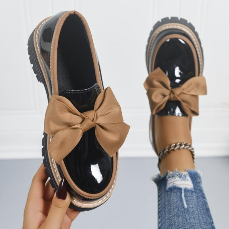 Beige slip-on loafers with black lining, bow detail, and chunky tread sole