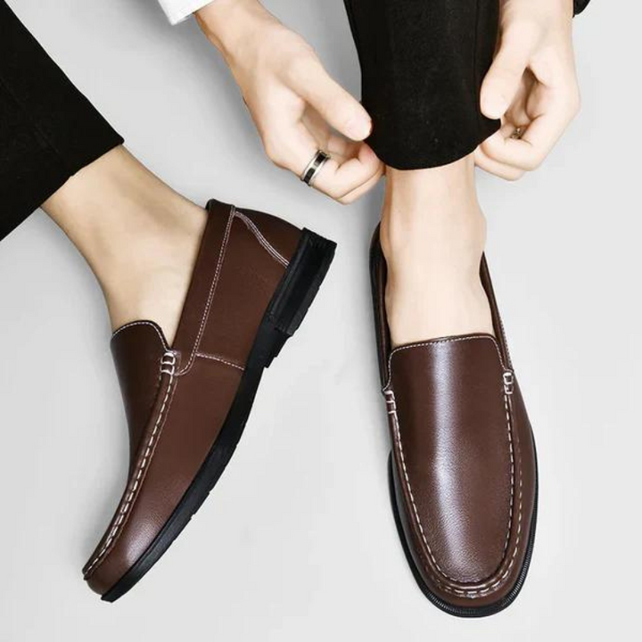 Brown leather penny loafers held by hands, metal bar detail and stitched trim