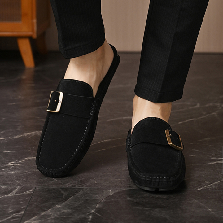 Black suede loafers with gold buckle strap, rear quarter view on feet