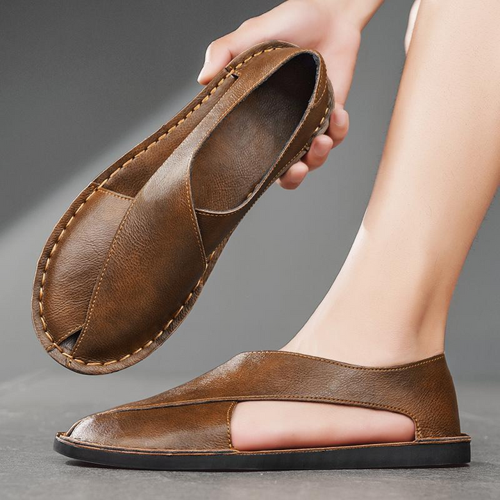 Brown leather open-toe slide sandal being held above a foot wearing the matching pair.