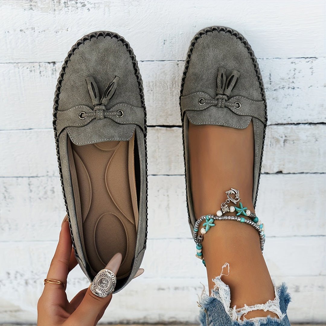 Gray suede tassel loafers with bow detail and scalloped moccasin stitching, flat sole.